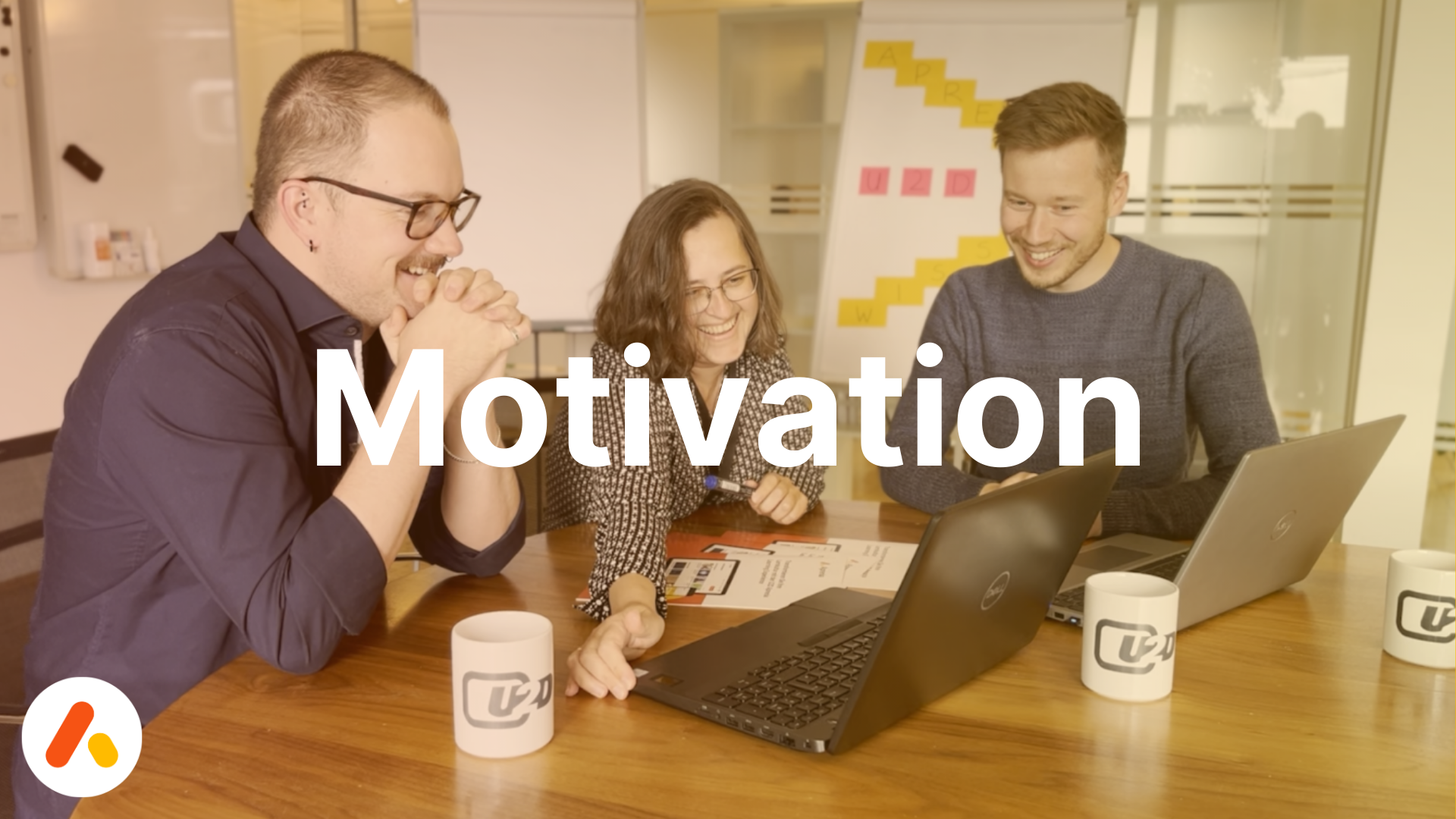 Drei Mitarbeiter*innen sitzen lachend an einem Tisch vor ihren Laptops, Schriftzug: Motivation